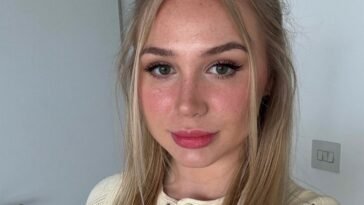Patty Peach, a young blonde woman with long straight hair, poses for a selfie indoors. She wears a cream knit button-up top and subtle glam makeup with pink lipstick and winged eyeliner. The soft natural lighting highlights her clear skin and fresh look.