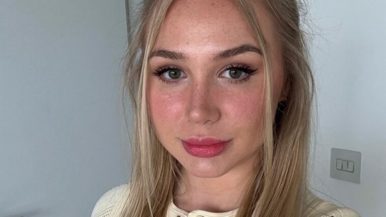 Patty Peach, a young blonde woman with long straight hair, poses for a selfie indoors. She wears a cream knit button-up top and subtle glam makeup with pink lipstick and winged eyeliner. The soft natural lighting highlights her clear skin and fresh look.