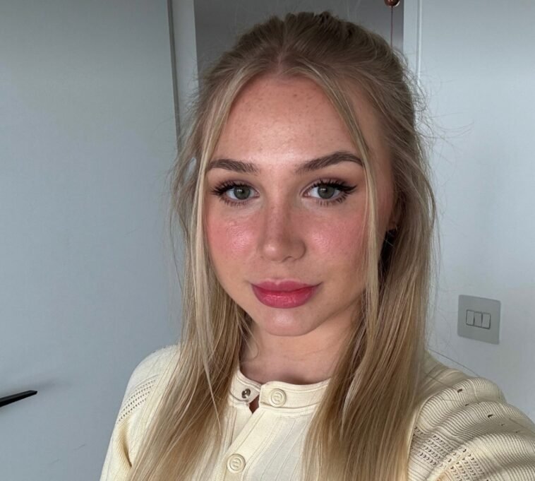 Patty Peach, a young blonde woman with long straight hair, poses for a selfie indoors. She wears a cream knit button-up top and subtle glam makeup with pink lipstick and winged eyeliner. The soft natural lighting highlights her clear skin and fresh look.