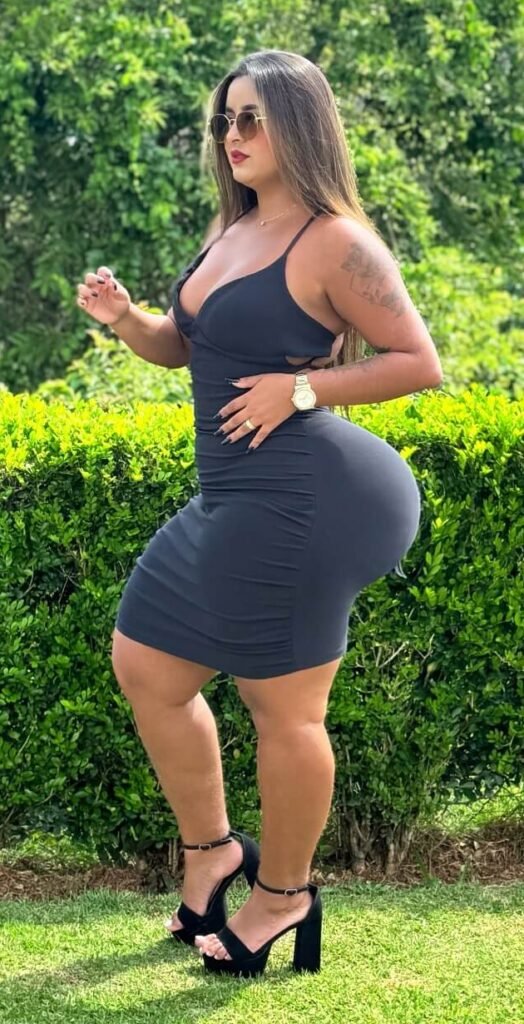 Dinalva Vasconcelos Wiki: Bio, Facts, Age, Height, Weight, Measurement, Photos; glamour Model 10 Dinalva Vasconcelos posing outdoors in a tight black dress and heels, showcasing her curvy figure with lush greenery in the background.