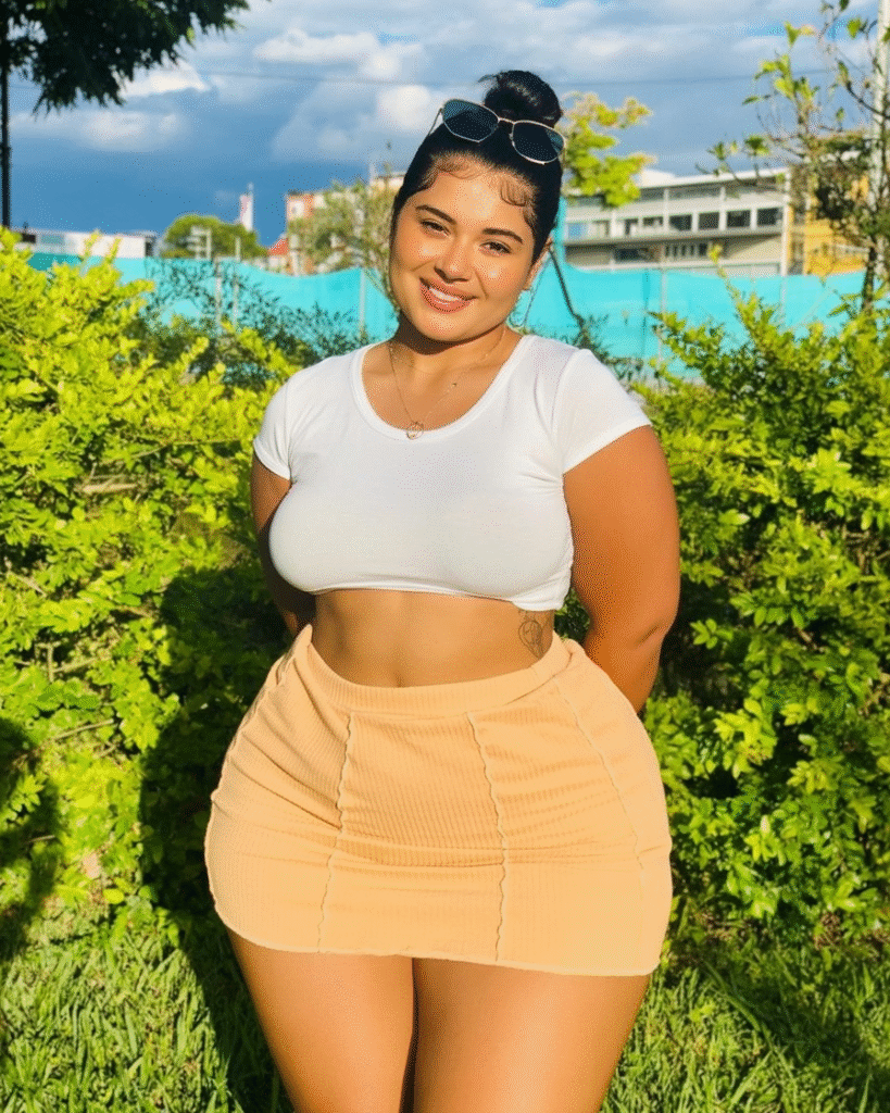 Who Is Angie Acosta? Biography, Wiki, Net Worth, Social Media, Measurements 10 Curvy Colombian fashion model Angie Acosta posing confidently outdoors in a white crop top and peach mini skirt, smiling under natural sunlight with green foliage and a blue sky in the background.