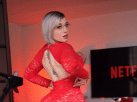 A stylish woman with short silver hair and glasses poses confidently in a red lace backless dress, showing a tattoo along her spine. She stands in a room lit with red lighting, next to a screen displaying the Netflix logo.