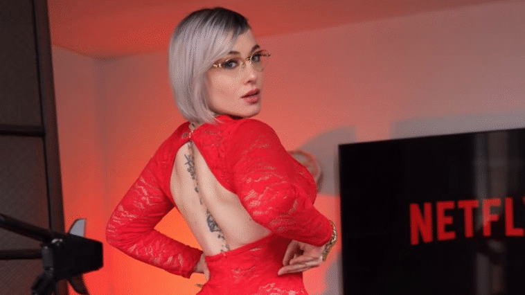 A stylish woman with short silver hair and glasses poses confidently in a red lace backless dress, showing a tattoo along her spine. She stands in a room lit with red lighting, next to a screen displaying the Netflix logo.