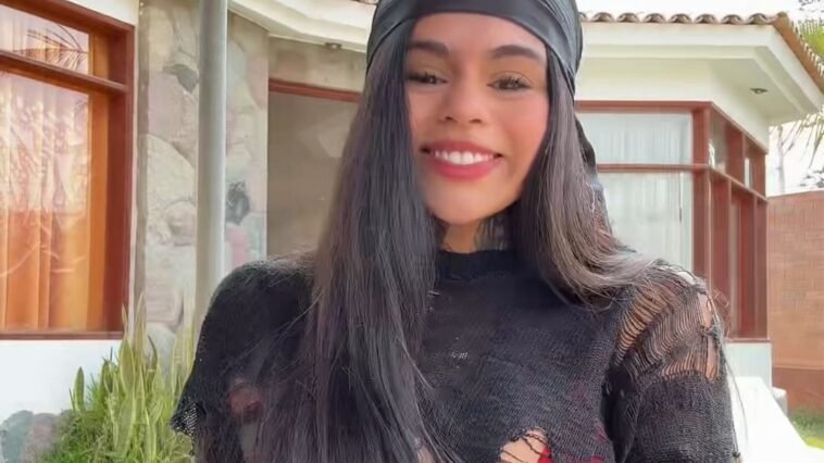 Victoria Hernández standing outdoors in front of a modern house, smiling brightly with a black headscarf and shredded black top.