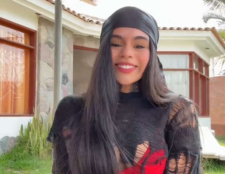 Victoria Hernández standing outdoors in front of a modern house, smiling brightly with a black headscarf and shredded black top.