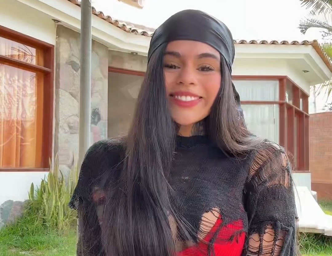 Victoria Hernández standing outdoors in front of a modern house, smiling brightly with a black headscarf and shredded black top.