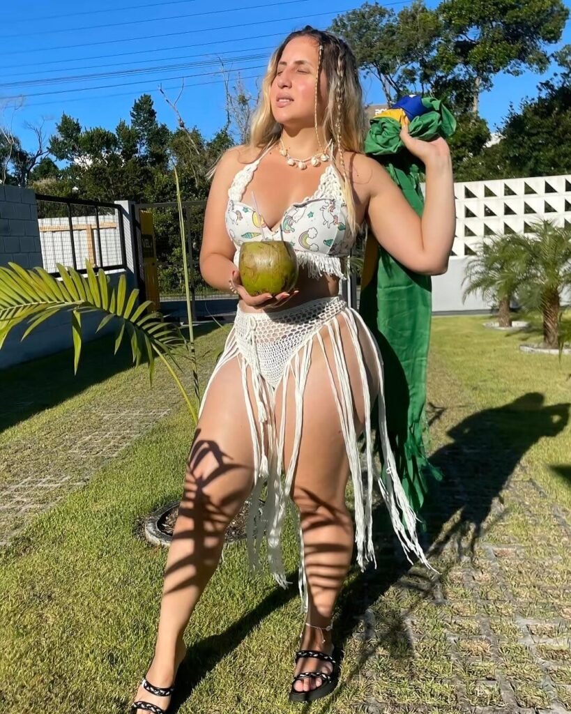Nathy Gallas Biography: Age, Height, Career, Net Worth, and Social Media Success 10 Nathy Gallas basking in the sun while holding a coconut, wearing a crochet skirt and printed bikini top, standing on a sunny lawn with a Brazilian flag.