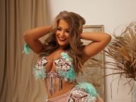 Viktorya Cherkashina kneels on sand in a pastel pink and turquoise feathered belly dance costume, smiling radiantly in a studio shoot.
