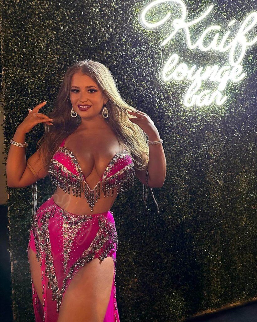 Viktorya Cherkashina: Age, Height, Weight, BIO, Family, Net Worth - Complete Guide 12 Viktorya Cherkashina poses smiling in front of a glowing “Kaif Lounge Bar” sign, wearing a sparkling fuchsia belly dance costume.