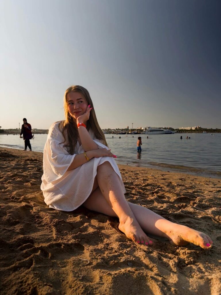 Viktorya Cherkashina: Age, Height, Weight, BIO, Family, Net Worth - Complete Guide 10 Viktorya Cherkashina sits barefoot on a golden beach at sunset, wearing a white wrap dress and smiling softly toward the horizon.
