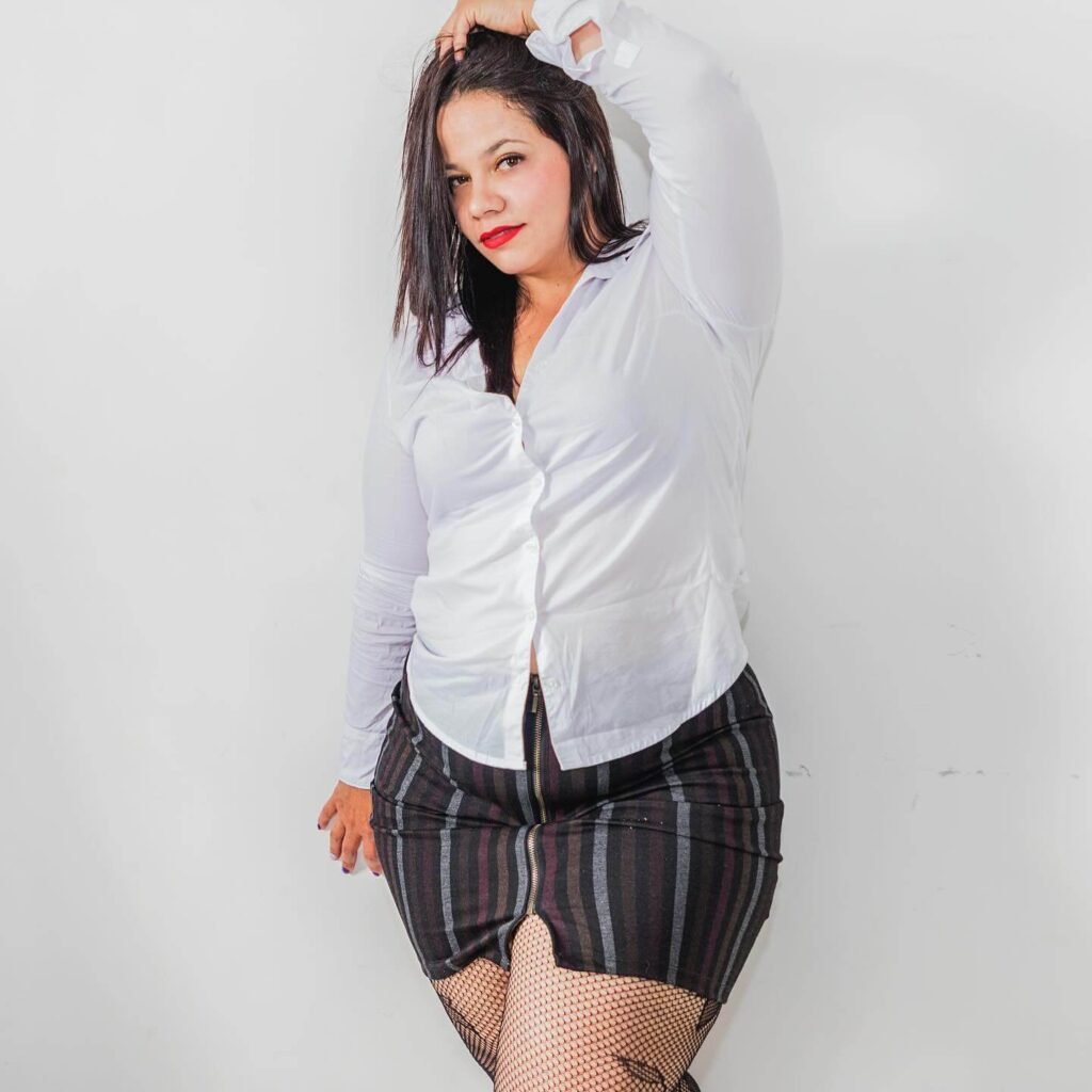Colombian plus-size model Yurley Rúa posing in a white button-up shirt and striped skirt with fishnet stockings, hand in hair, confident fashion pose.