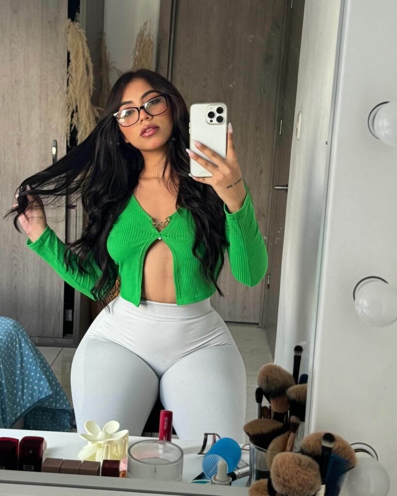 Angel Lopez taking a mirror selfie in a green cropped cardigan and white leggings, showing her long dark hair and glasses.