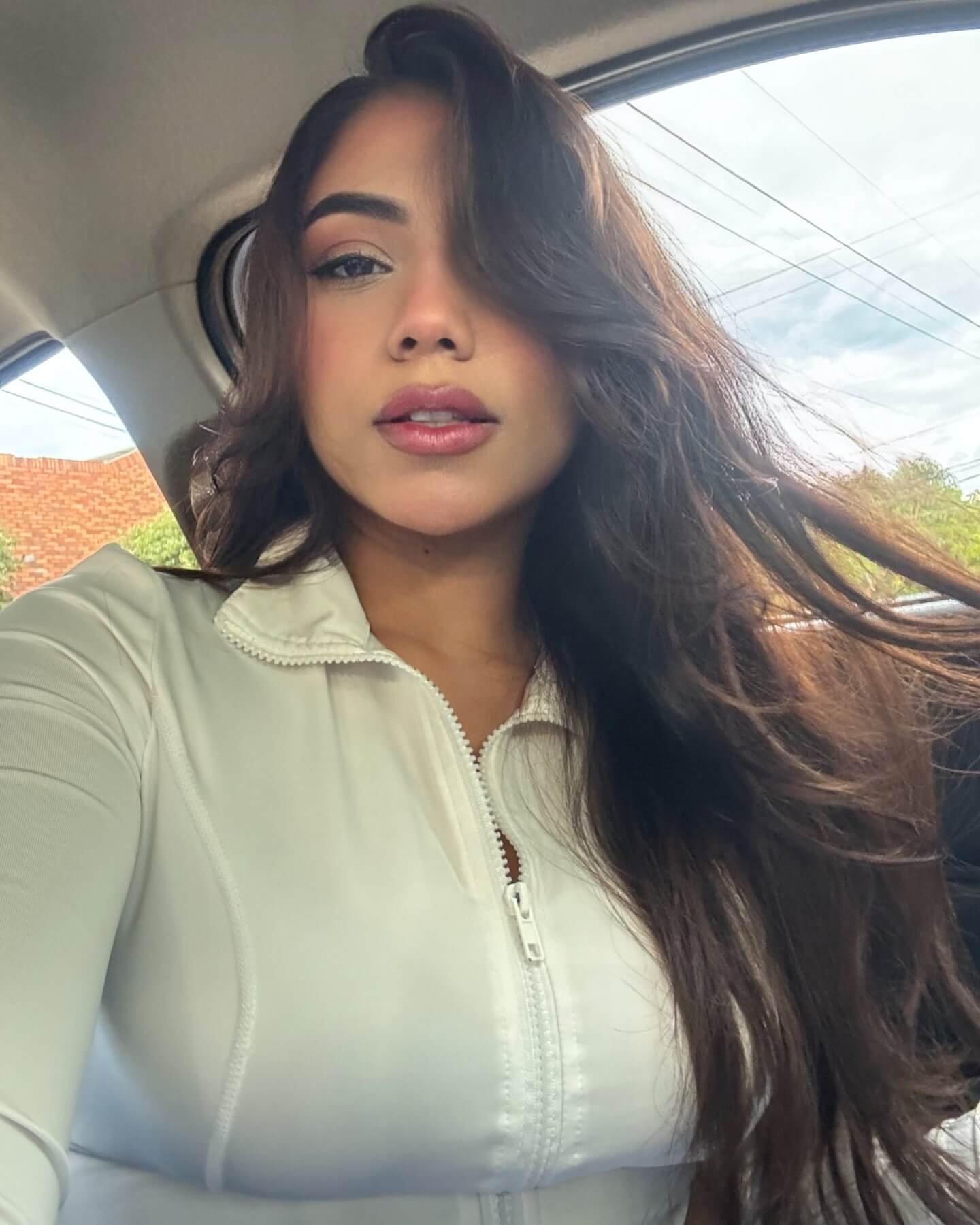 Close-up selfie of Angel Lopez wearing a white zip-up top with loose hair in a car.