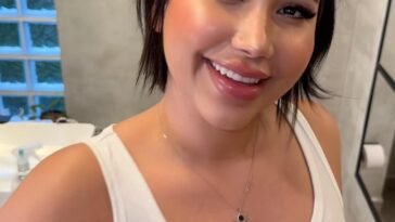 Smiling Anna Beggion in a white ribbed tank top with short dark hair, standing in a bright modern bathroom.
