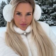 Close-up of Jolie Becker outdoors in winter, wearing white earmuffs and a white zip-up jacket with snow-covered trees in the background.