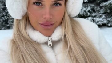 Close-up of Jolie Becker outdoors in winter, wearing white earmuffs and a white zip-up jacket with snow-covered trees in the background.