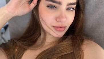 Close-up selfie of Laura Sahar with natural makeup, long brown hair, and a floral dress under natural sunlight.