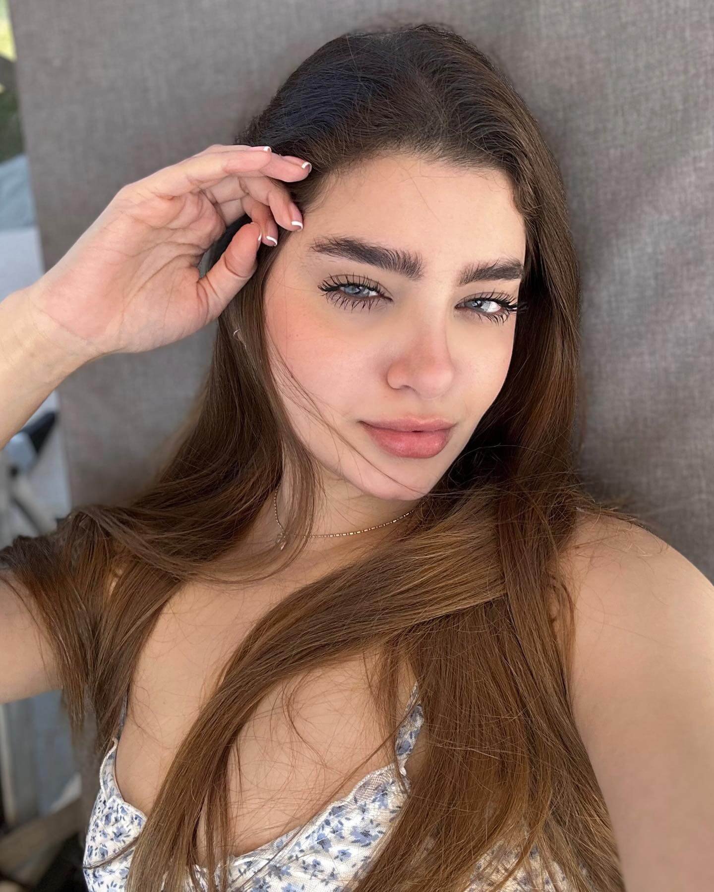 Close-up selfie of Laura Sahar with natural makeup, long brown hair, and a floral dress under natural sunlight.