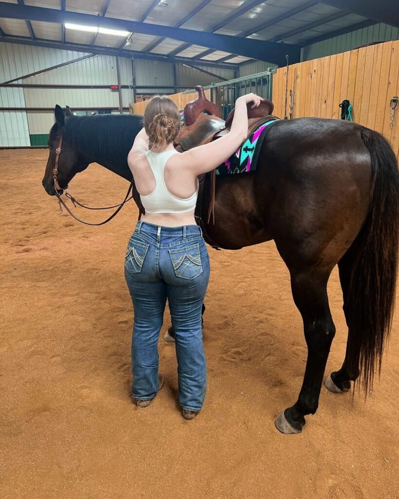 Shawnee Youngblood standing with her back to the camera, holding a brown horse in an indoor riding arena while wearing jeans and a white crop top.